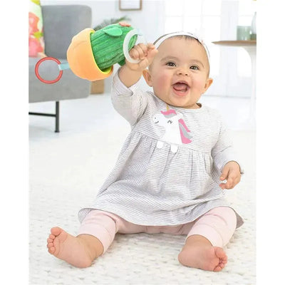 Baby wearing a grey striped dress with a unicorn graphic joyfully holds a Skip Hop Cactus Jitter Hanging Toy.
