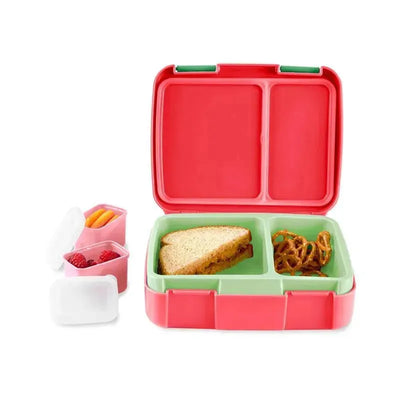 Bento lunch box in strawberry color with sandwich, pretzels, and small containers of fruit and vegetables.