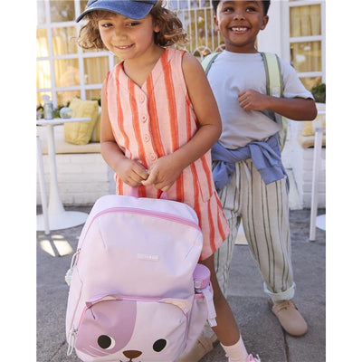 Toddler girl wearing a striped dress holds a pink Skip Hop cat backpack with padded straps, smiling at the camera.
