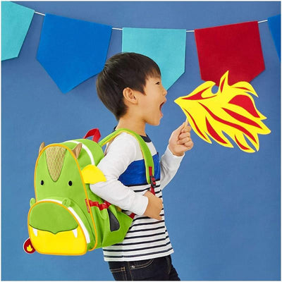 A boy wearing a striped shirt and a green dinosaur-themed Skip Hop backpack makes a roaring gesture with a fire prop.
