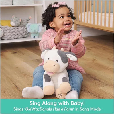 Animated plush cow, Buttermilk, sits on the lap of a smiling child who is clapping, with vibrant nursery decor in the background.