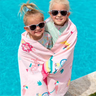 Two young girls, wearing sunglasses, smile while wrapped in a colorful beach towel by a pool.