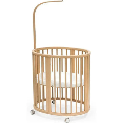 Natural wood crib with a round design and a white mattress, featuring wheels for mobility and a movable arm for toys.