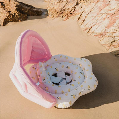 Baby float in the shape of a flower, featuring a pink canopy and floral patterns, set on a sandy beach.