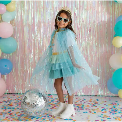 A child models the Sweet Wink Aqua Star Cape dress, wearing a light blue cape, ruffled dress, and decorative sunglasses, beside a disco ball and colorful balloons.
