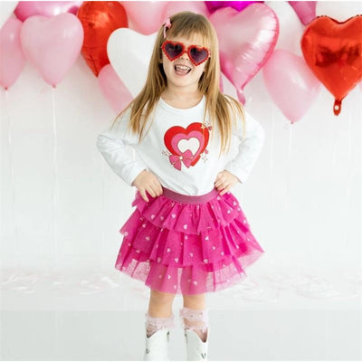 A girl smiles and poses in a white long-sleeve shirt with a heart design and a pink layered tutu skirt, surrounded by heart-shaped balloons.
