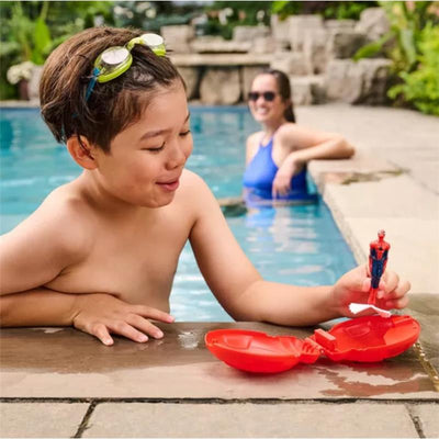A young boy with swimming goggles plays with a red Swimways Marvel Spider-Man Dive N Surprise toy by a pool.