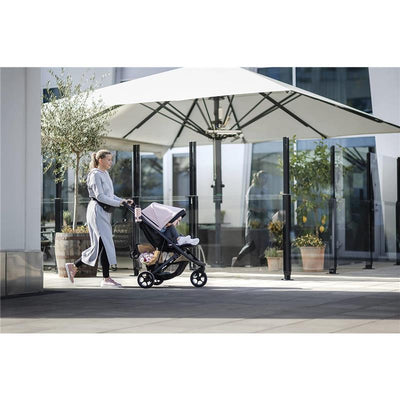 A woman in a grey outfit walks with a stroller featuring a pink seat, carrying a child and a bag of flowers outdoors.