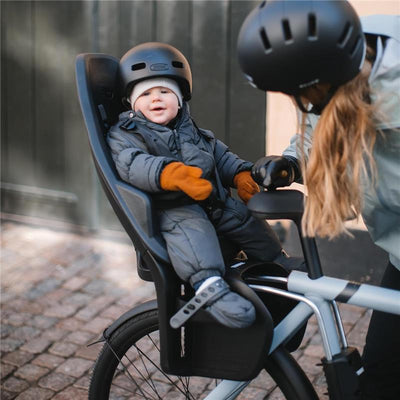 Thule Yepp 2 Maxi Rack Mount child bike seat on a bicycle with a smiling toddler wearing a helmet and gloves, assisted by a woman.