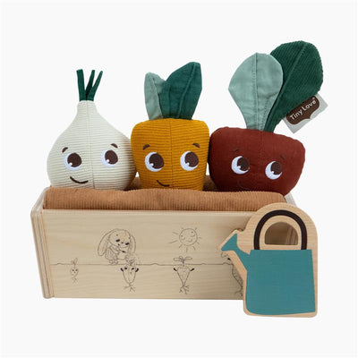 Soft toy vegetables in a wooden planter box, featuring an onion, carrot, and beet with smiling faces and a watering can toy.