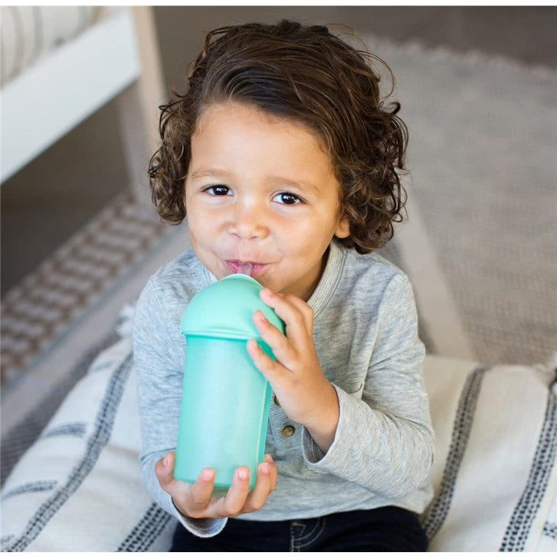 Child with curly hair sipping from a mint-colored Boon SWIG Silicone Straw Cup, sitting on a patterned blanket.