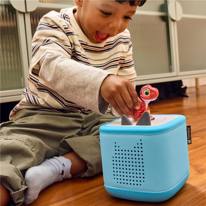 Young child joyfully places a red dinosaur figurine on a blue Tonies audio box while sitting on the floor.