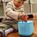 Young child joyfully places a red dinosaur figurine on a blue Tonies audio box while sitting on the floor.