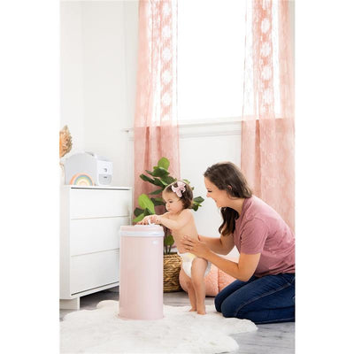 A mother helps her toddler use a blush pink Ubbi Steel Diaper Pail in a bright, softly decorated room.