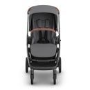Uppababy Kona stroller with a gray and charcoal color scheme, featuring a padded seat, safety harness, and a brown handlebar.