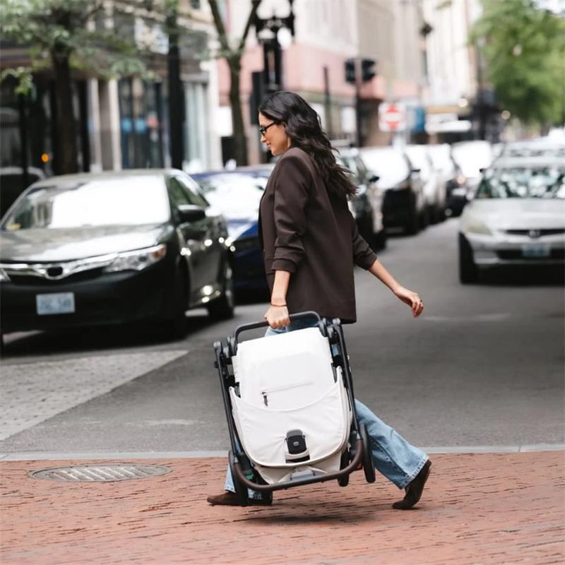 A woman walks along the street, carrying a folded Uppababy Kona stroller and bassinet bundle in Ada (Sandstone Mélange) color.