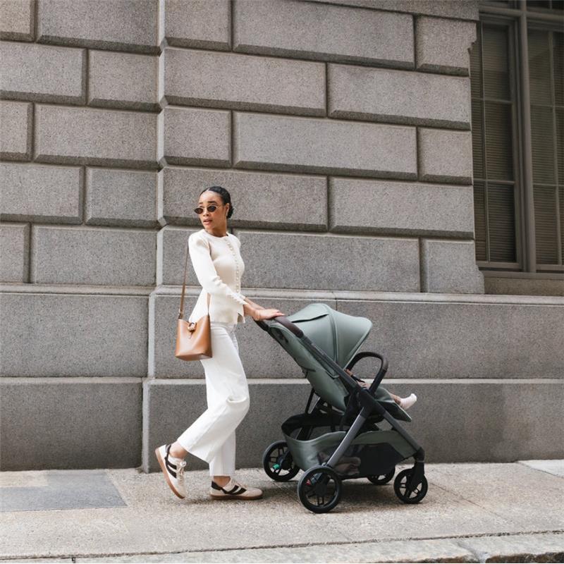 Woman in white outfit walks with Uppababy Kona stroller in James (black) against a textured building wall.