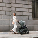 Woman in a stylish outfit walking with the Uppababy Kona stroller in James (Black) color against a stone wall.
