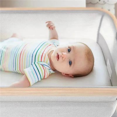 A baby in a striped onesie lays in a white bassinet with wooden accents, designed for safe sleeping.