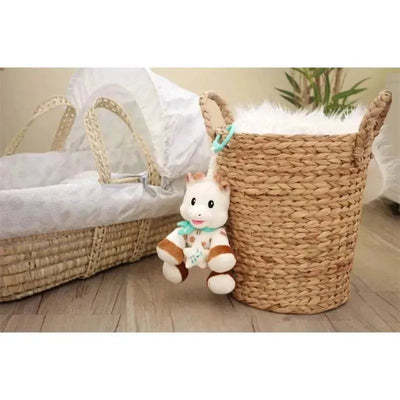 Plush Vulli Sophie La Girafe toy with musical box, hanging from a woven basket beside a baby bassinet.