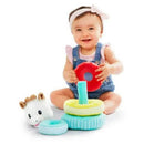 Smiling baby in denim overalls holds a red ring, playing with a colorful stacking toy featuring a plush giraffe.