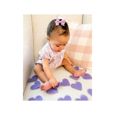 Light orchid grosgrain hair bow on a no-slip clip worn by a baby girl sitting on a couch with purple heart shapes.