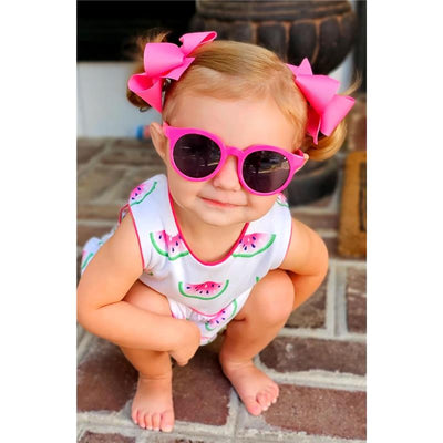 Hot pink grosgrain hair bows in pigtails, on a baby wearing watermelon-patterned outfit and sunglasses, crouching on brick.