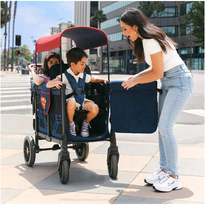 WonderFold W2 Luxe Pro double stroller wagon, Superman design, with two children seated; an adult is attaching a storage bag.