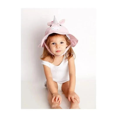 Toddler wearing a pink alicorn sunhat with ears and a horn, seated and looking at the camera.